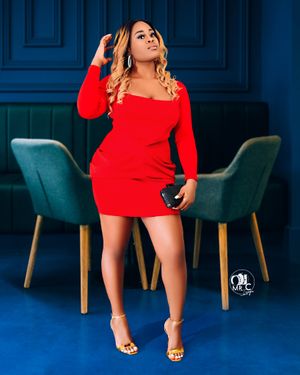 A woman in a fitted red dress strikes a pose between two teal chairs against a dark blue wall.