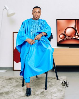 A person wearing a bright blue traditional agbada sits elegantly on a chair in a studio setting.