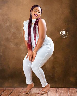 Studio portrait showing person in white outfit with burgundy braids striking a playful pose on wooden floor.