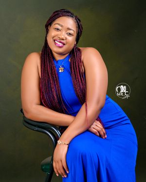 Professional portrait of someone in a royal blue dress with burgundy braids sitting in a chair against a dark background.