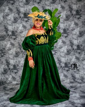 Person in dramatic emerald green velvet gown with matching hat and tropical foliage backdrop.