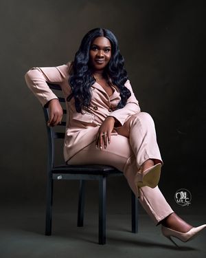 Elegant pose in blush pink suit lounging on black chair against dark background.
