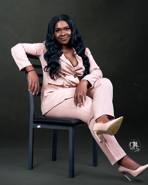 Professional portrait in pink pantsuit seated on black chair against dark backdrop.