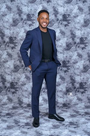 Man in tailored navy suit and black shirt poses against textured grey studio background.