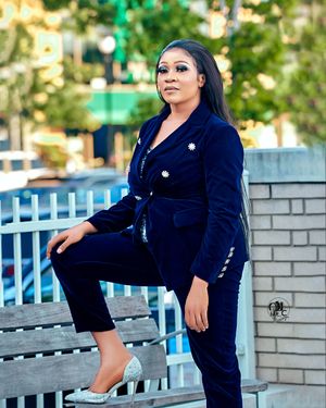 Woman in navy velvet pantsuit poses on park bench with urban background.