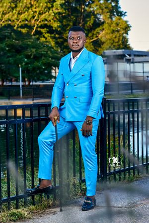Man in bright blue double-breasted suit poses confidently against outdoor railing at sunset.