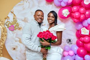 Joyful couple posing with red roses against pink balloon arch and white backdrop.