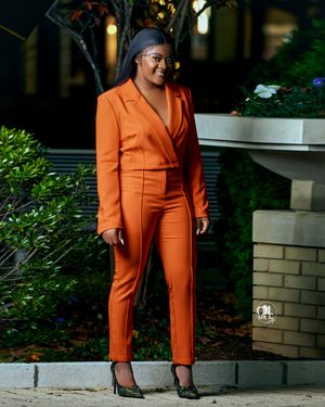 Professional portrait of person wearing bright orange fitted pantsuit against garden backdrop.
