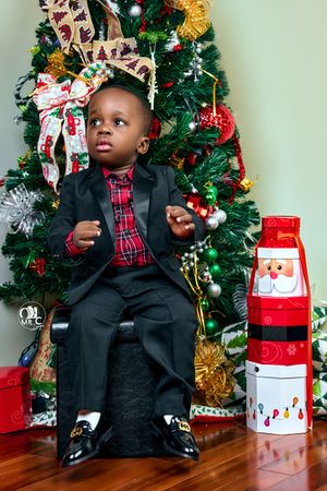 Young child in formal suit sitting by Christmas tree with Santa decoration.