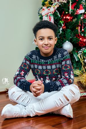 Child wearing festive holiday sweater sitting cross-legged by Christmas tree.