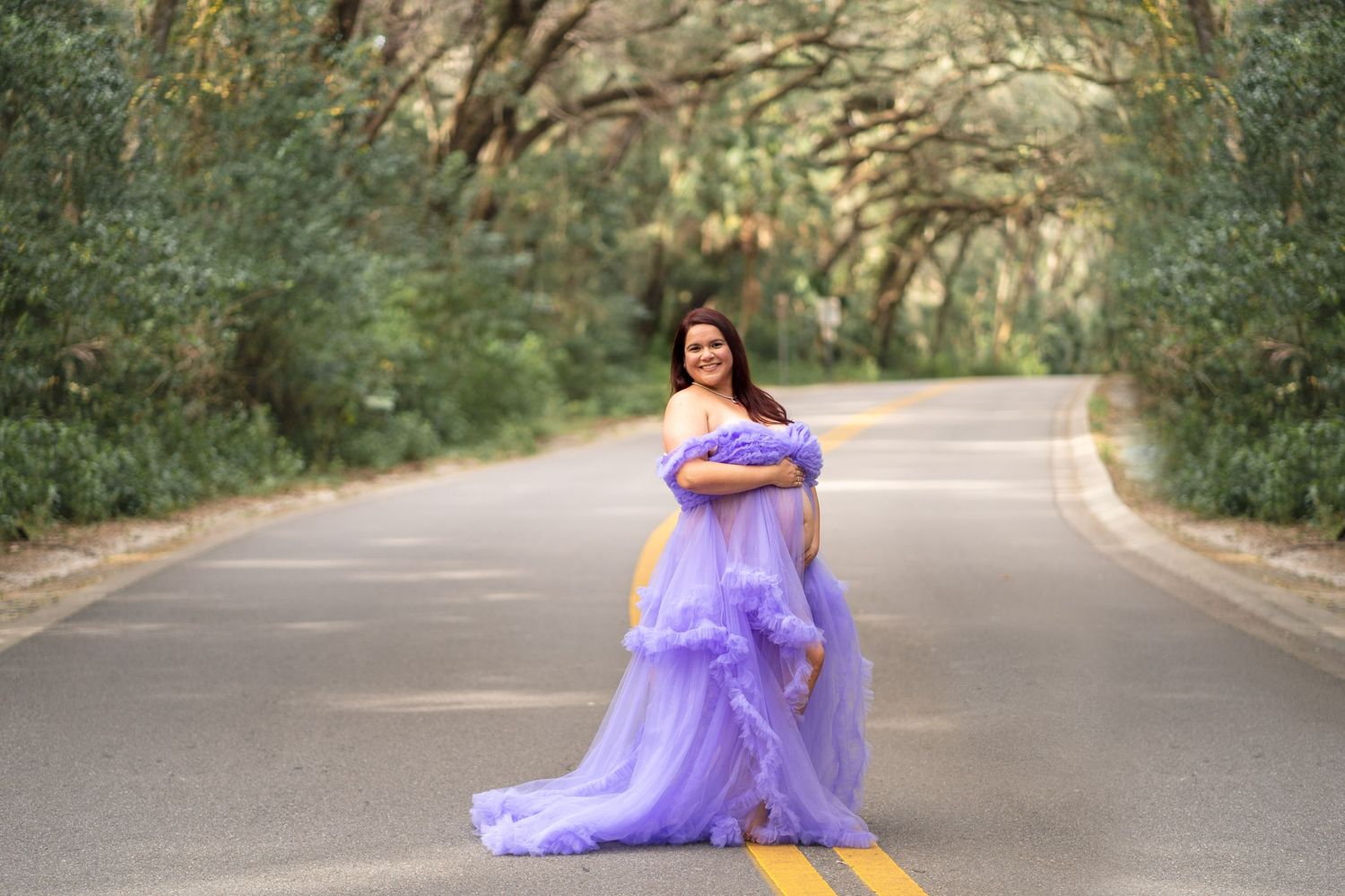 Person in flowing purple gown poses on tree-lined road.