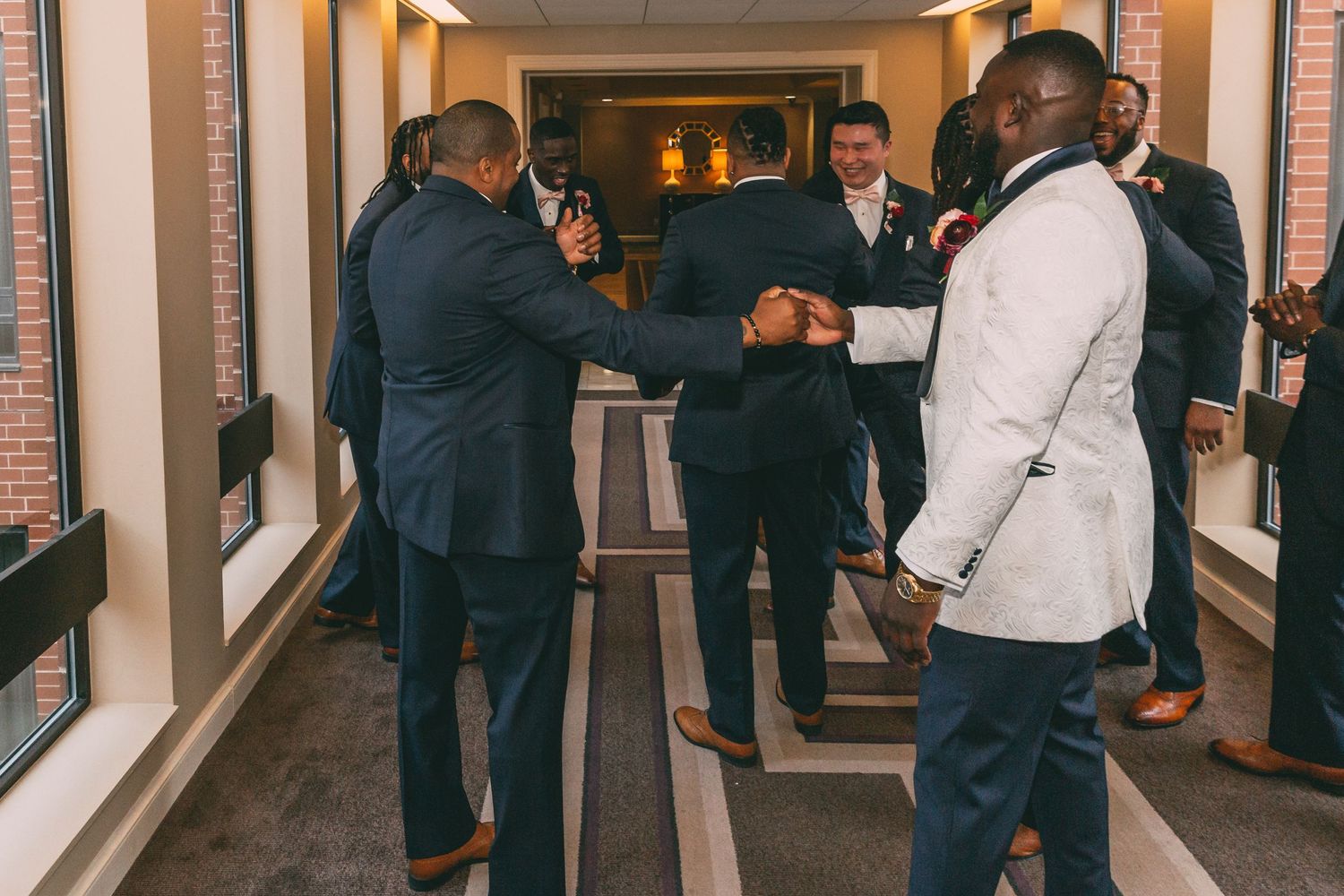 Groomsmen in navy suits greet and congratulate each other with handshakes in a hallway before the wedding ceremony.