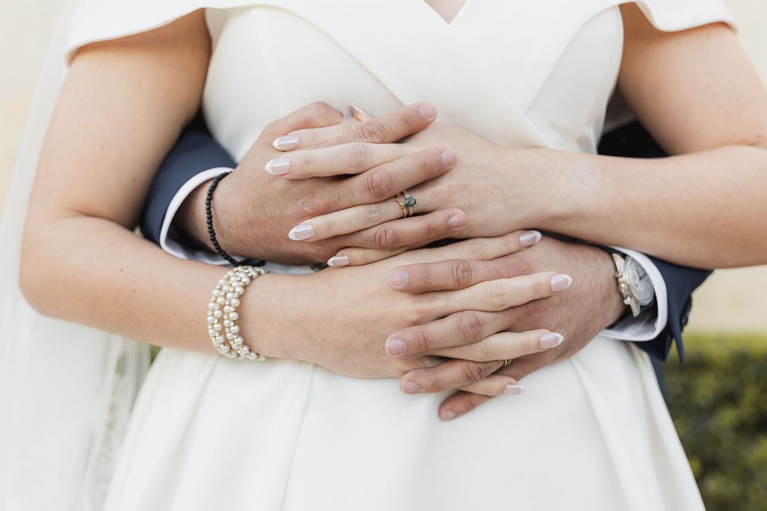 Close-up of intertwined hands with wedding rings and pearl bracelet against white dress.