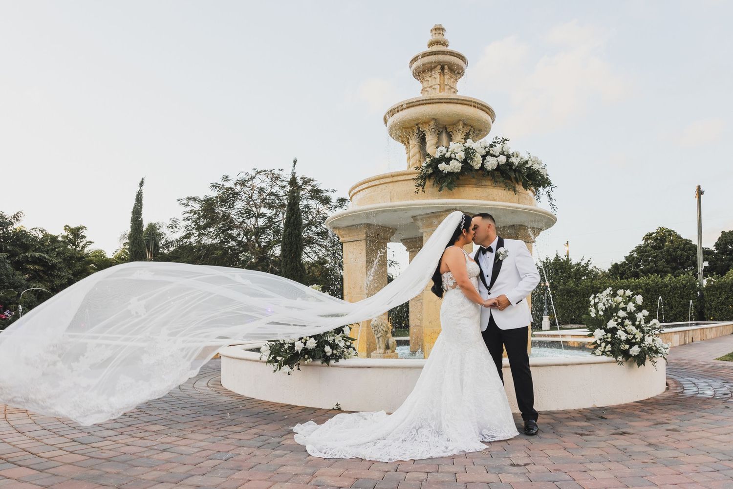 Dramatic wedding portrait with flowing veil at ornate fountain during golden hour.