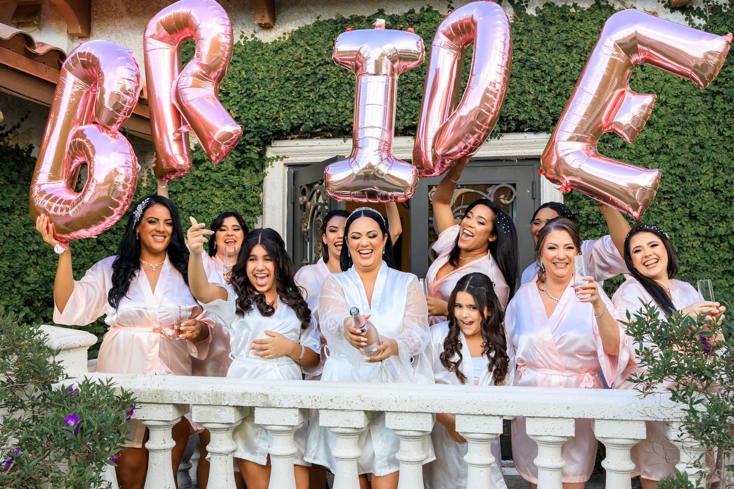 Celebratory bridal party gathering with rose gold letter balloons spelling BRIDE against greenery backdrop.