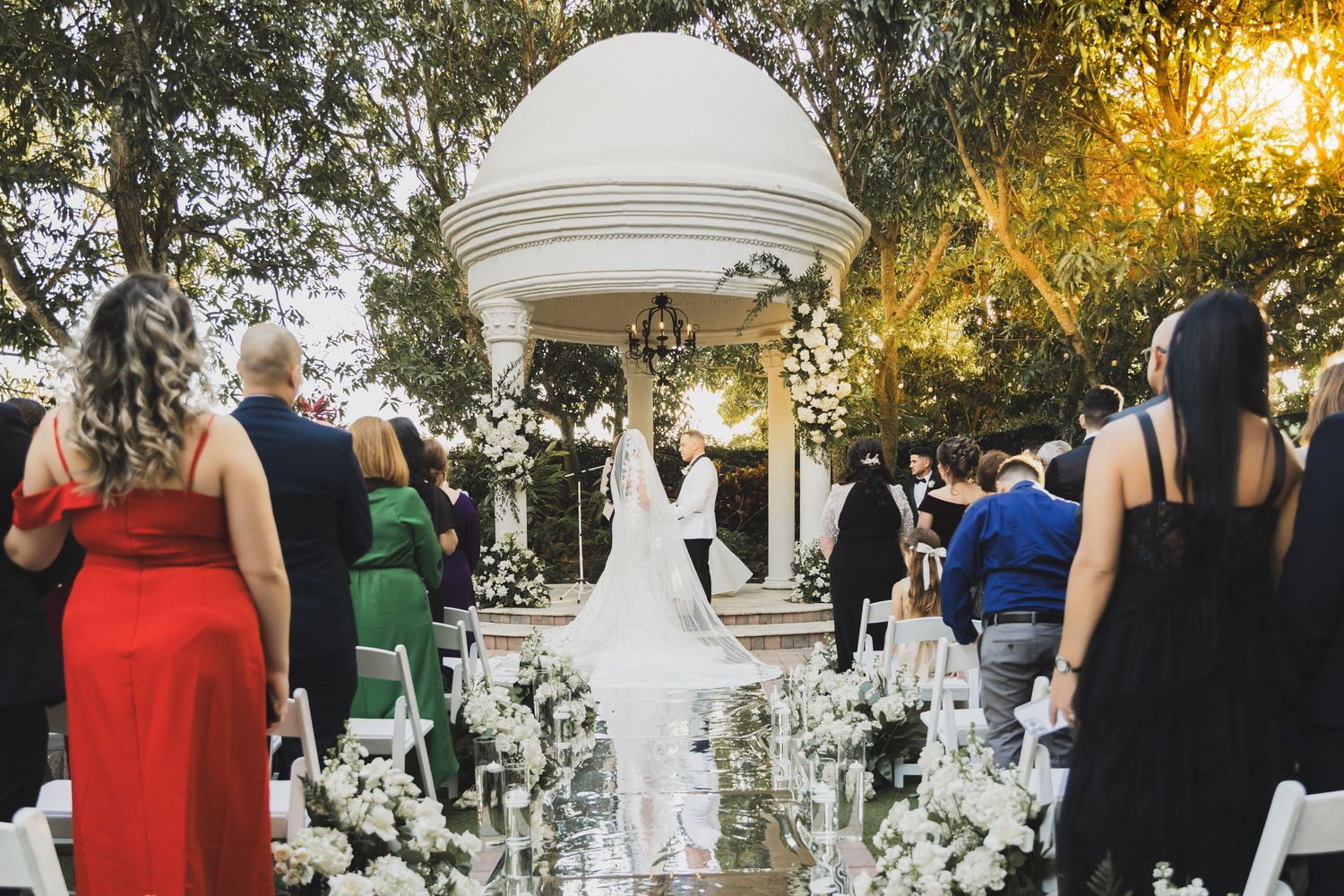 Elegant outdoor wedding ceremony at white gazebo surrounded by floral arrangements and seated guests.