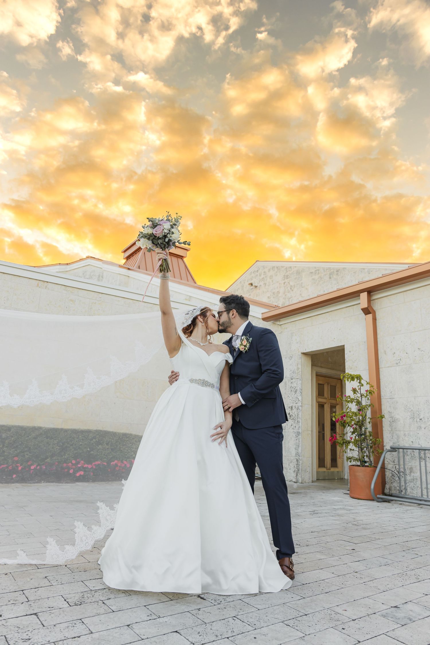 A wedding couple celebrates against a dramatic sunset sky on a terrace.