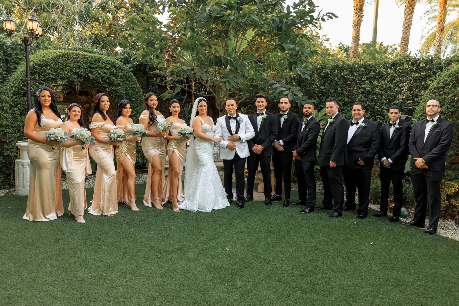 Wedding party portrait with bridesmaids in champagne dresses and groomsmen in black tuxedos on lawn.