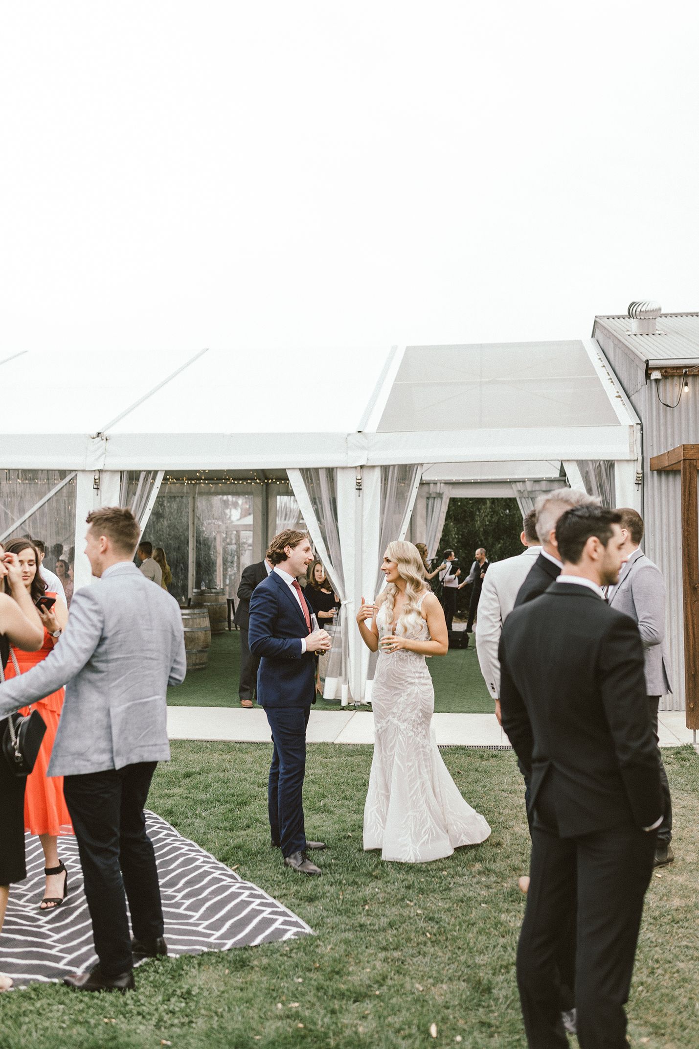 Outdoor wedding reception under white tent with guests mingling on grass.