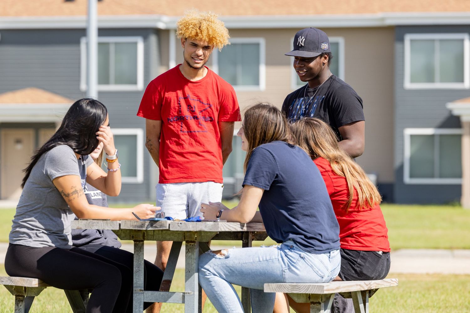 Students gather around an outdoor picnic table on a sunny day at a college campus.