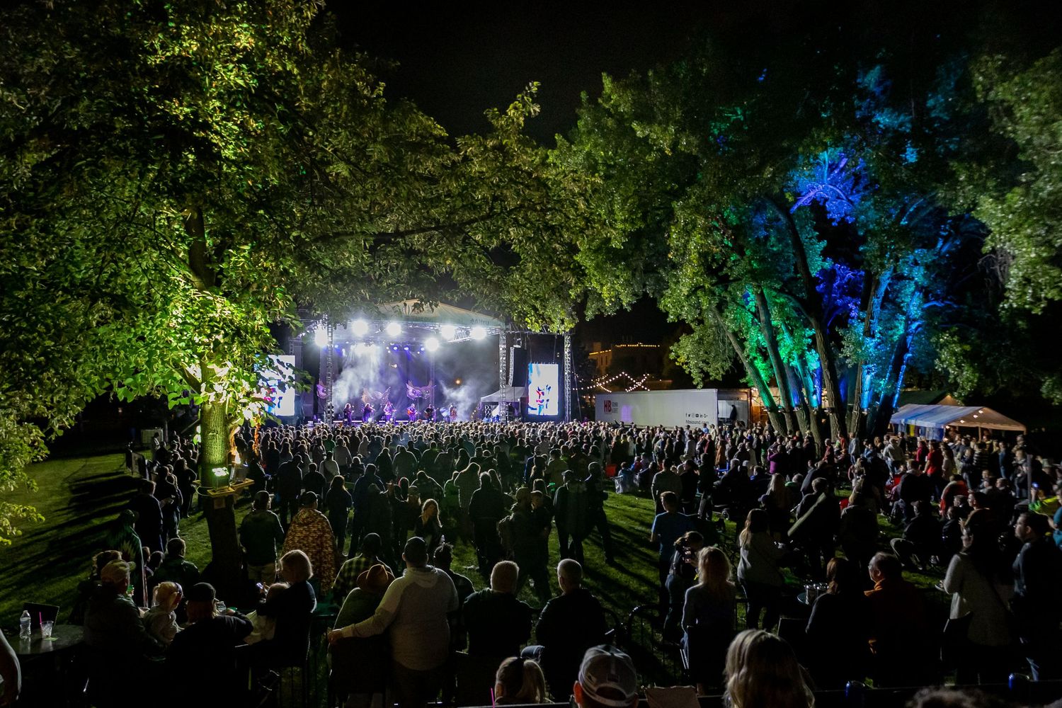 Large festival crowd gathered outdoors at night with trees lit up in green and purple.