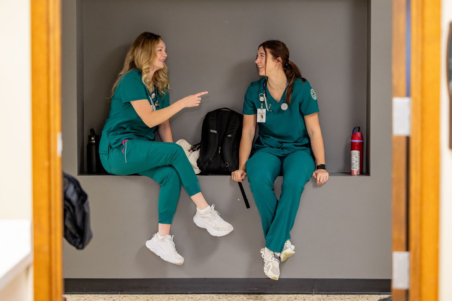 Healthcare workers in teal scrubs having a conversation while sitting in a hallway.