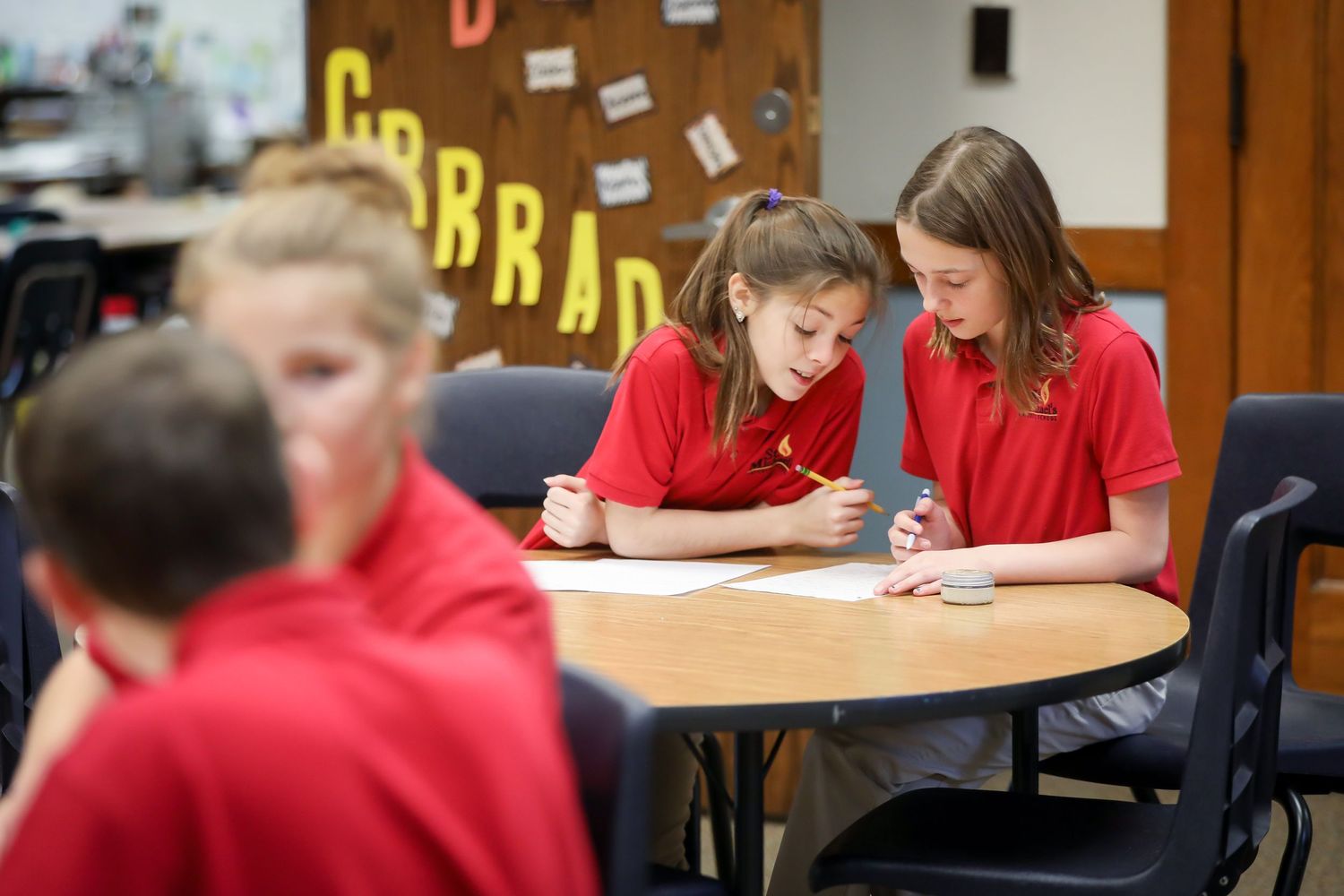 Students in red uniforms working together at round tables in classroom setting.