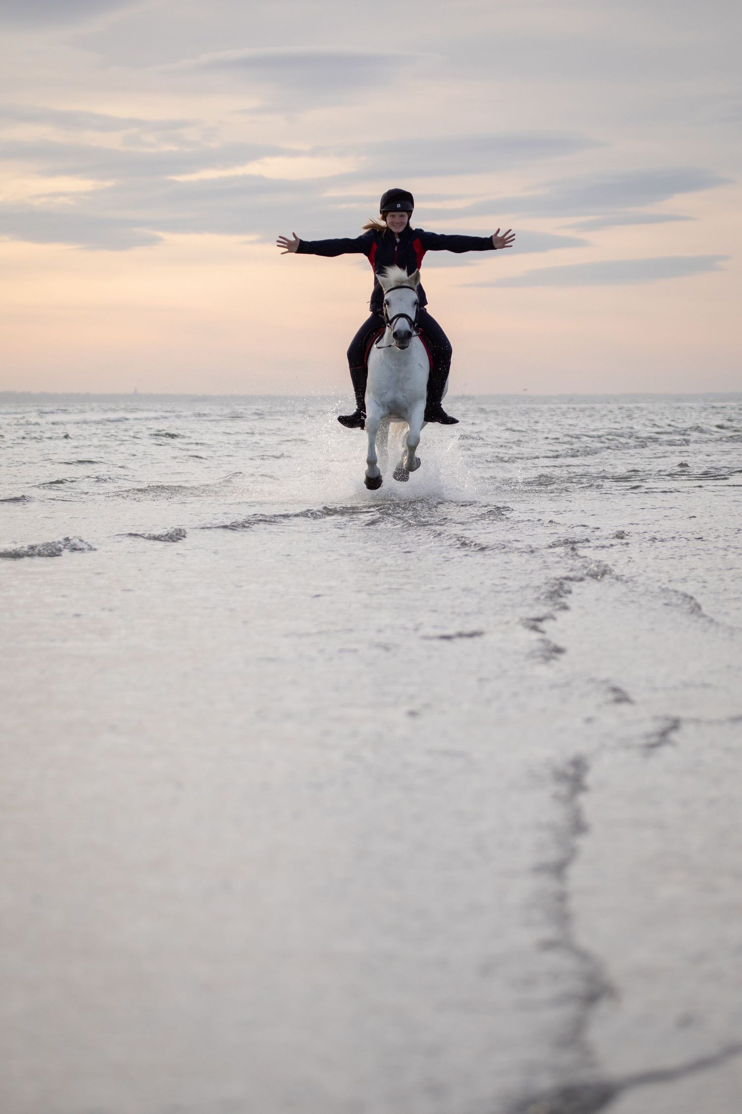 A person rides horseback with arms spread wide along the ocean shore at sunset.