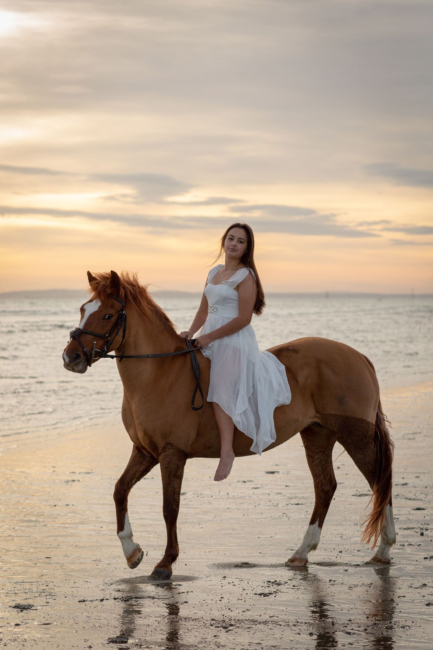 A person in white dress rides a chestnut horse along the beach at sunset.