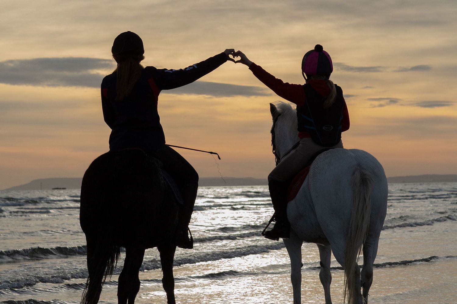Two riders on horseback create an arch with their arms during sunset on the beach.