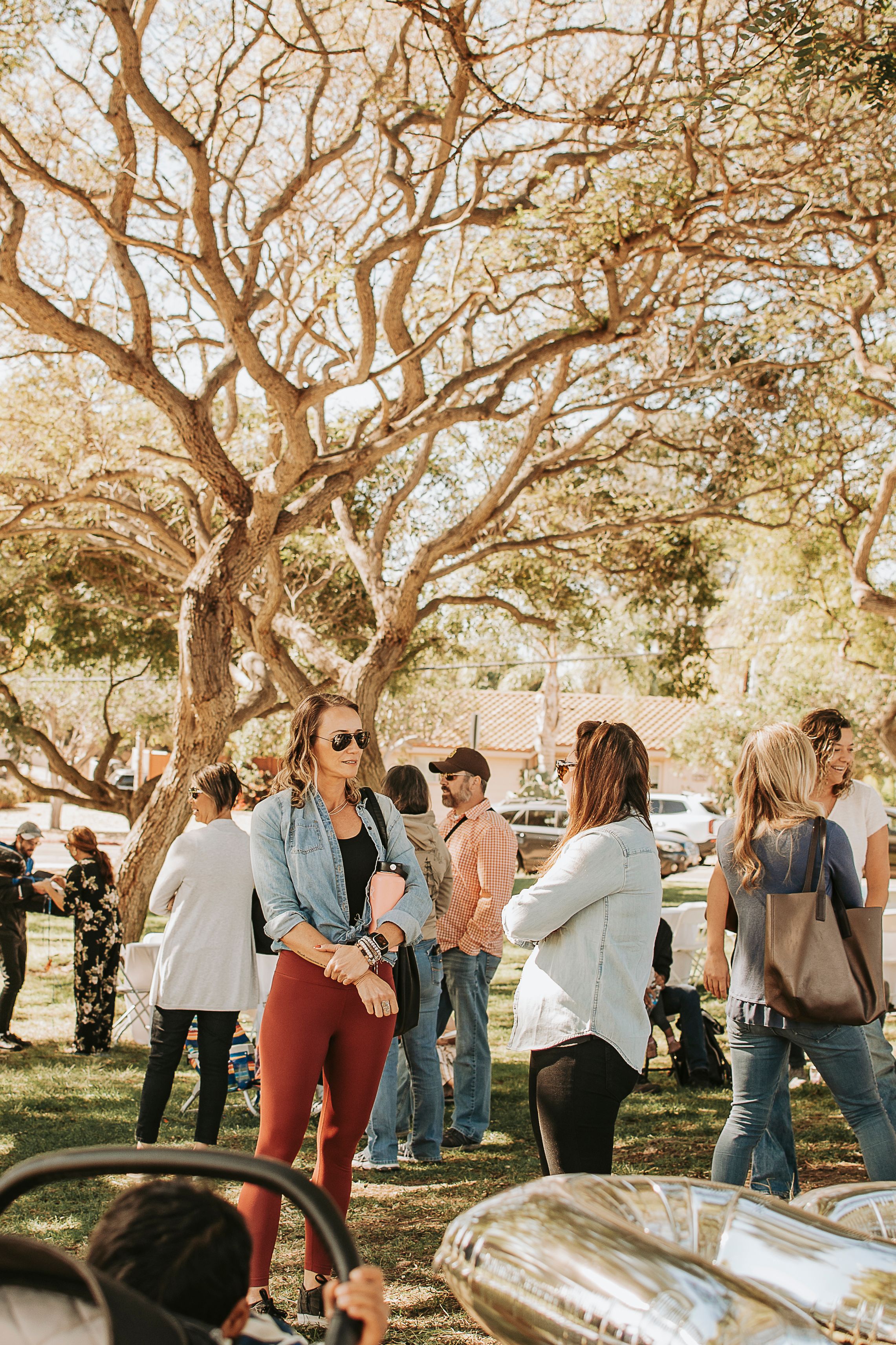 People gathered under large branching trees at an outdoor social event.