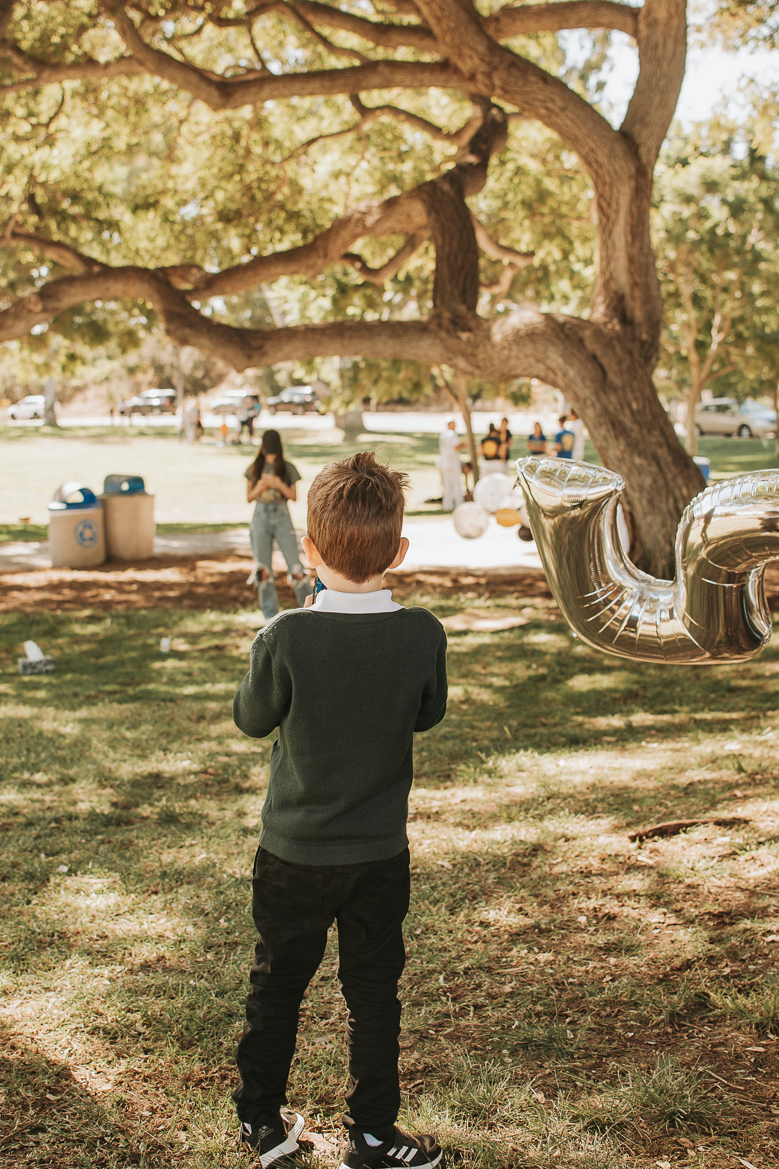 A child in a dark sweater stands watching a celebration under a large tree.