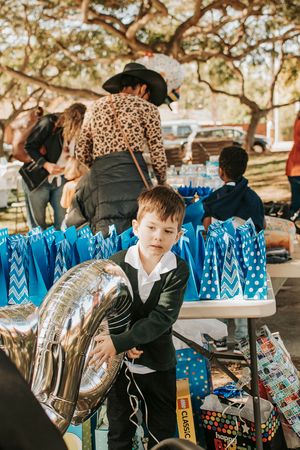 A child next to blue gift bags and silver balloons at an outdoor party.