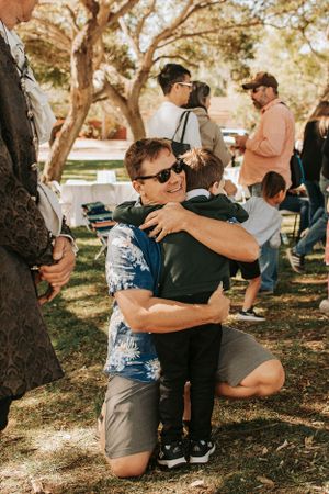 People gathered at an outdoor event under large shady trees.
