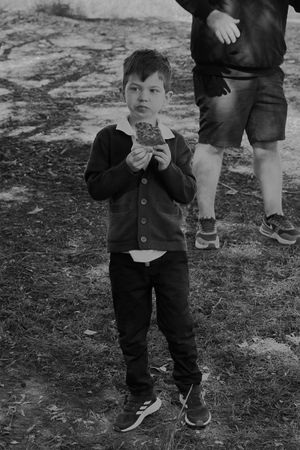 Young child dressed in dark clothing standing outdoors looking at a slice of pizza.
