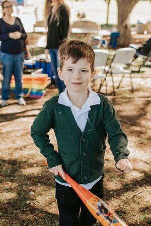 Young child in a green cardigan holds an orange toy while standing in a park.