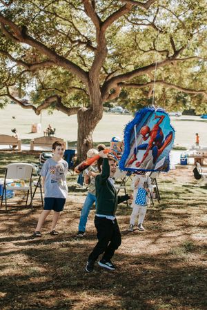 Children playing with colorful pinatas at an outdoor party under large oak trees.