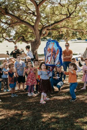 Large group of children gathered for a pinata party under shaded trees.