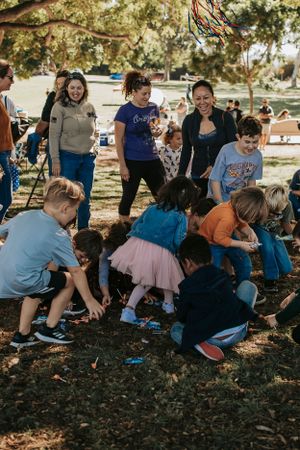 Large group enjoying a lively pinata party under oak trees in a park setting.