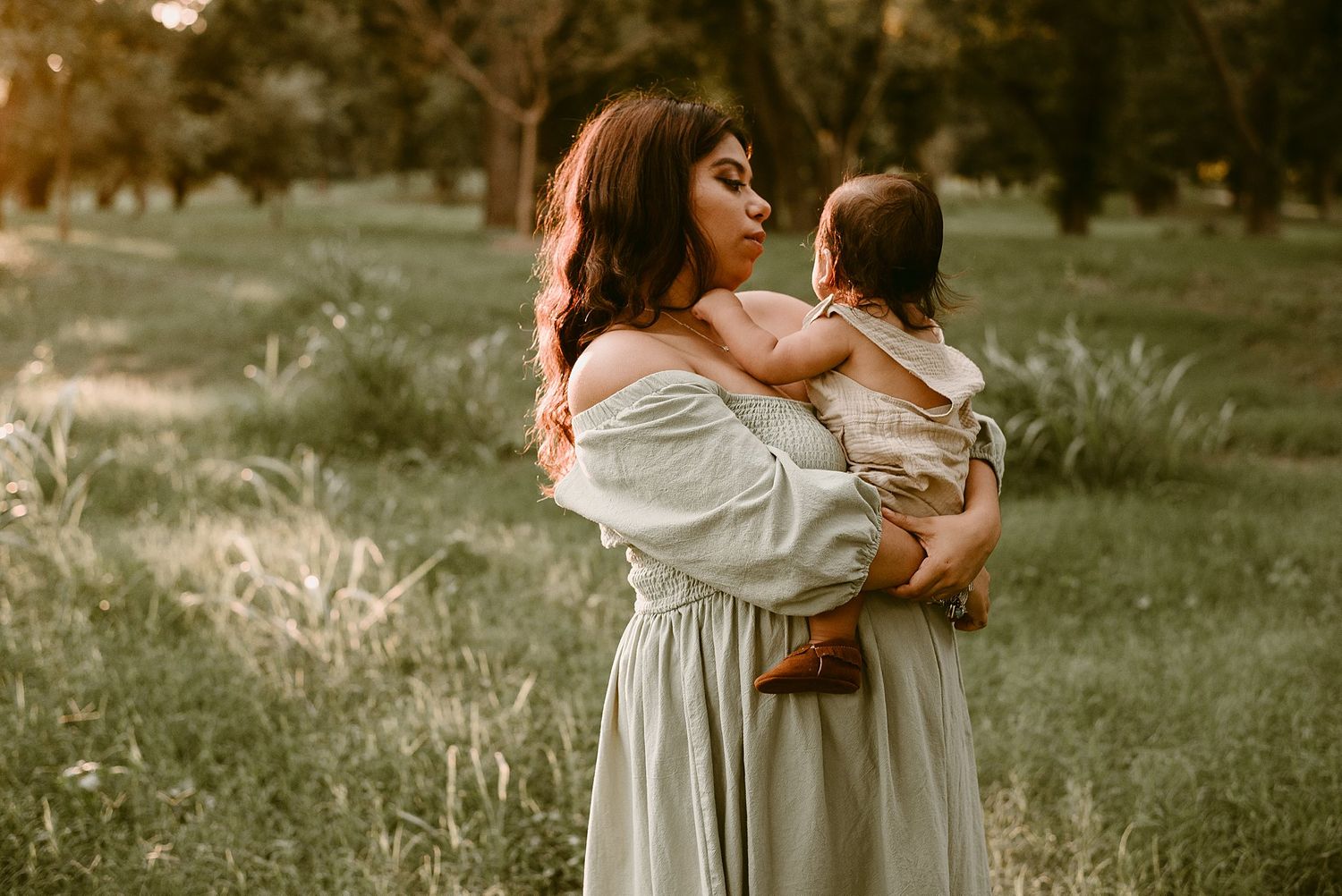 A woman in a flowing dress holds a baby against her shoulder in a sunlit park setting.