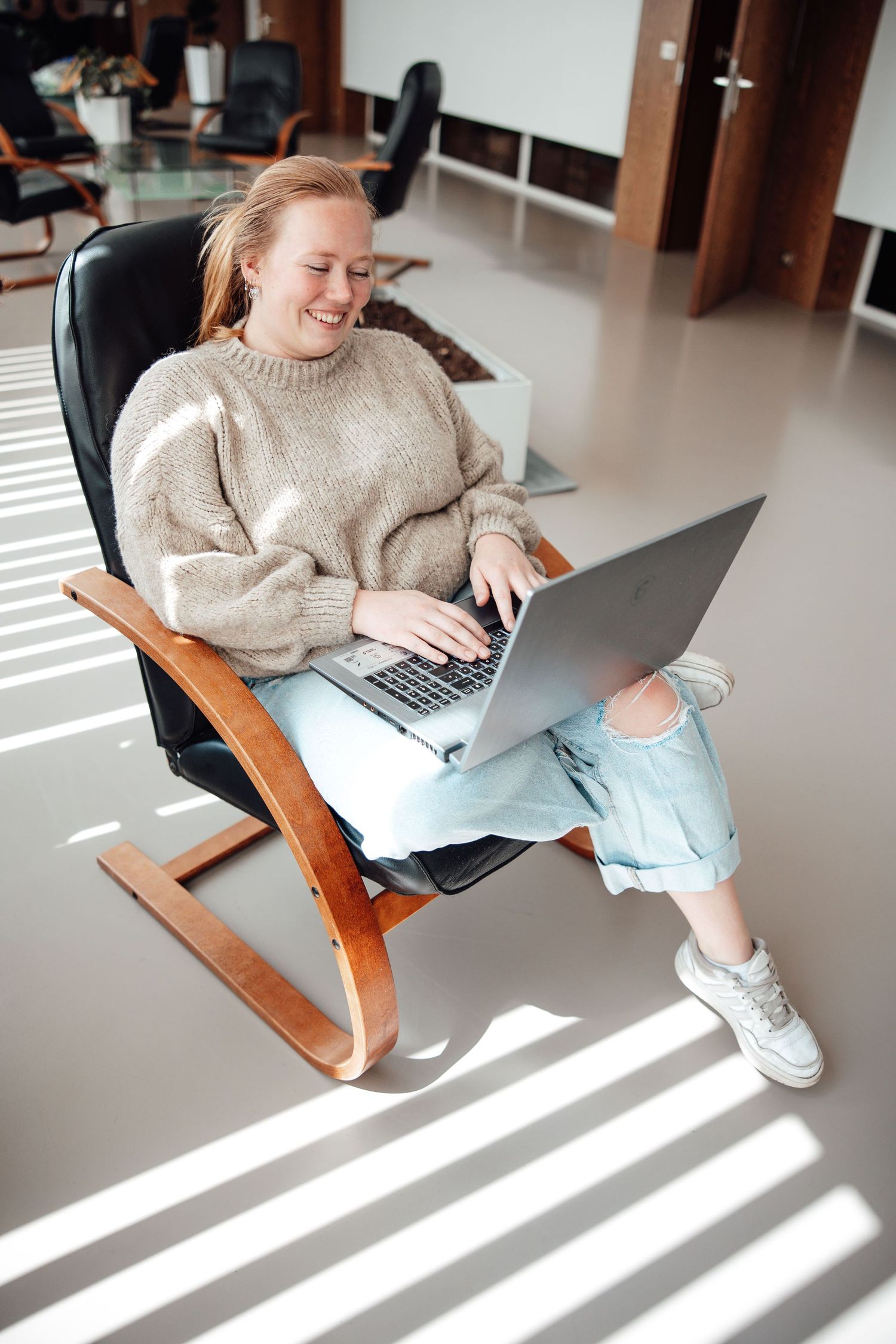 Person in cozy sweater works on laptop while relaxing in modern wooden rocking chair.