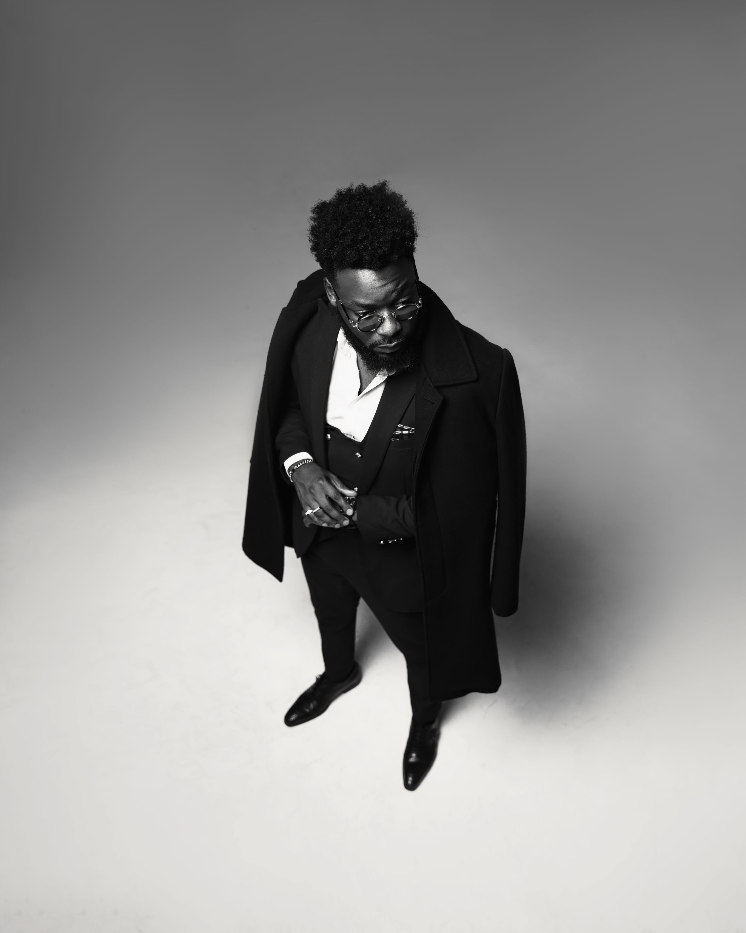 Stylish black and white portrait of someone in an overcoat looking down against a plain backdrop.