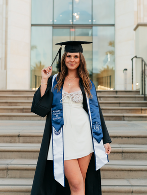 Graduate in cap and gown with blue sash poses on steps of academic building.