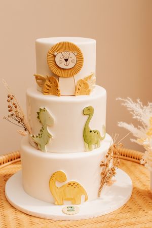 Three-tiered white cake decorated with cute dinosaurs and lion designs in pastel colors.