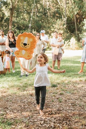 A child in a white dress runs joyfully with an orange lion piñata at an outdoor party.