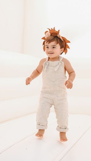 Toddler in beige overalls and lion headpiece stands smiling against white background.