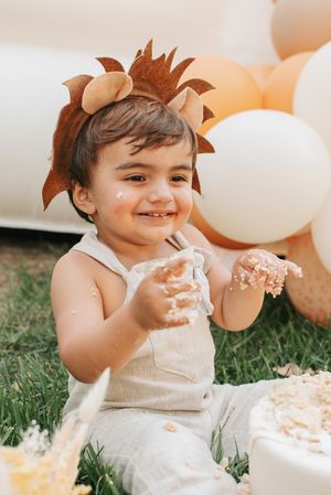 Toddler wearing lion headpiece enjoys smashing cake while sitting on grass with balloons in background.