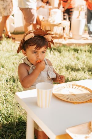 Child wearing brown lion headpiece eating at white party table outdoors.