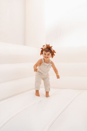 Toddler in white overalls and autumn leaf crown plays on white background.