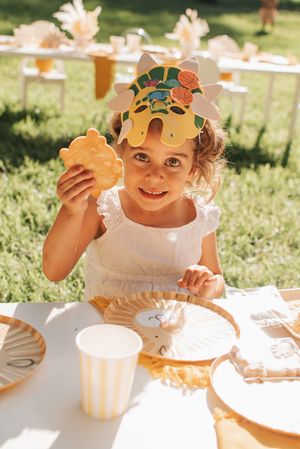Child wearing white dress and leaf crown enjoys treats at an outdoor autumn party.
